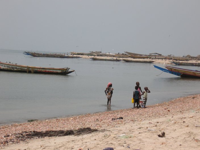 Vier Kinder stehen am Strand. Im Hintergrund erkennt man traditionelle, kleine Holzboote, die im Wasser liegen. 