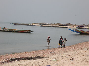Vier Kinder stehen am Strand. Im Hintergrund erkennt man traditionelle, kleine Holzboote, die im Wasser liegen. 