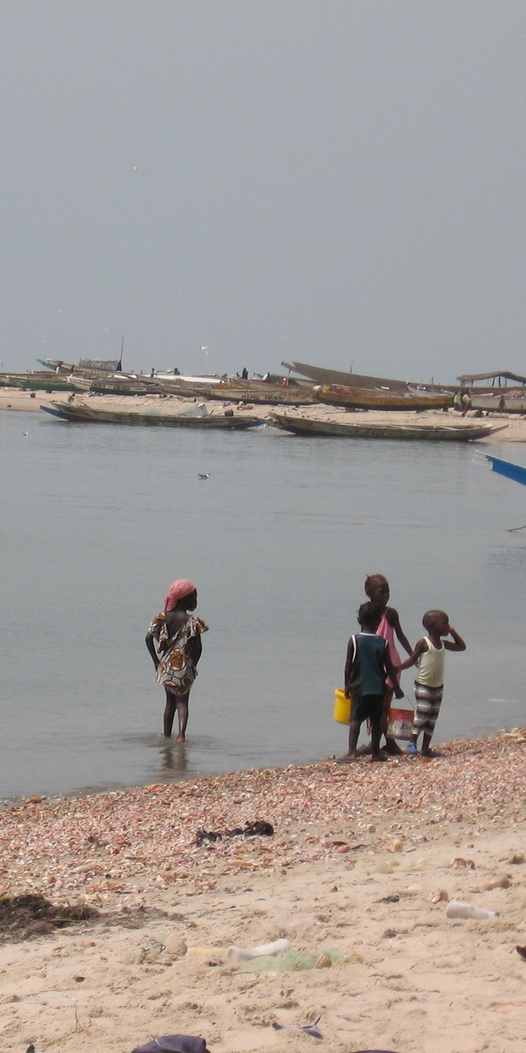 Vier Kinder stehen am Strand. Im Hintergrund erkennt man traditionelle, kleine Holzboote, die im Wasser liegen. 