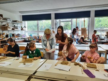 Workshop am Burghardtgymnasium in Buchen Schülerinnen aus der Ukraine sitzen in einer Klasse und kneten Ton im Rahmen eines Workshops. Eine Lehrerin steht in der Mitte und gibt Erklärungen.