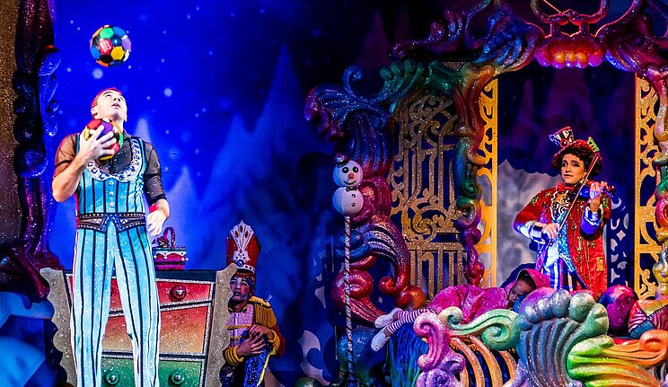 Das Bild zeigte eine Theaterszene vor bunter Theaterkulisse. Links im Bild steht ein Mann in einem glitzerenden Streifenkostum, der jongliert. Er schaut nach oben. Rechts steht in einem bunten Torbogen eine weitere Person mit einem Hut, die Geige spielt. In der Mitte steht eine bunte Kommode, hinter der sich eine Person kauernd versteckt.