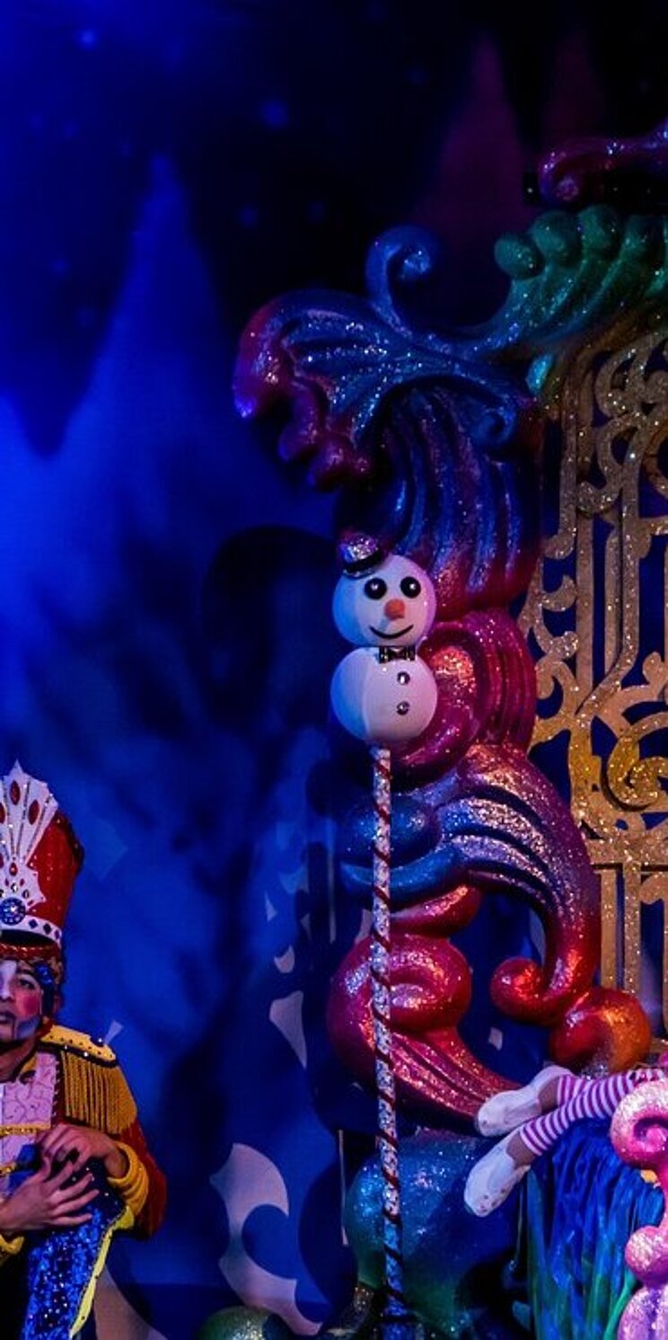 Das Bild zeigte eine Theaterszene vor bunter Theaterkulisse. Links im Bild steht ein Mann in einem glitzerenden Streifenkostum, der jongliert. Er schaut nach oben. Rechts steht in einem bunten Torbogen eine weitere Person mit einem Hut, die Geige spielt. In der Mitte steht eine bunte Kommode, hinter der sich eine Person kauernd versteckt.