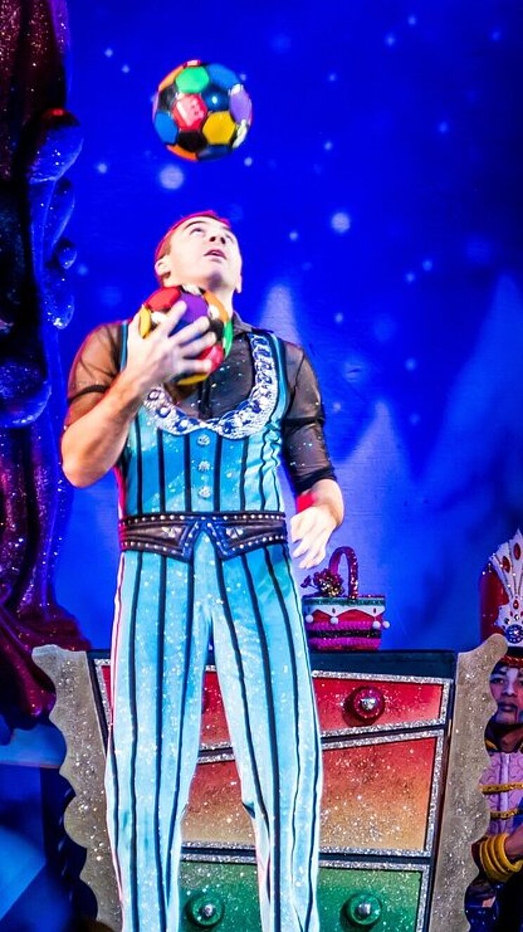 Das Bild zeigte eine Theaterszene vor bunter Theaterkulisse. Links im Bild steht ein Mann in einem glitzerenden Streifenkostum, der jongliert. Er schaut nach oben. Rechts steht in einem bunten Torbogen eine weitere Person mit einem Hut, die Geige spielt. In der Mitte steht eine bunte Kommode, hinter der sich eine Person kauernd versteckt.