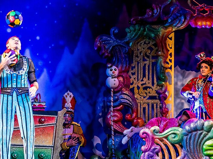 Das Bild zeigte eine Theaterszene vor bunter Theaterkulisse. Links im Bild steht ein Mann in einem glitzerenden Streifenkostum, der jongliert. Er schaut nach oben. Rechts steht in einem bunten Torbogen eine weitere Person mit einem Hut, die Geige spielt. In der Mitte steht eine bunte Kommode, hinter der sich eine Person kauernd versteckt.