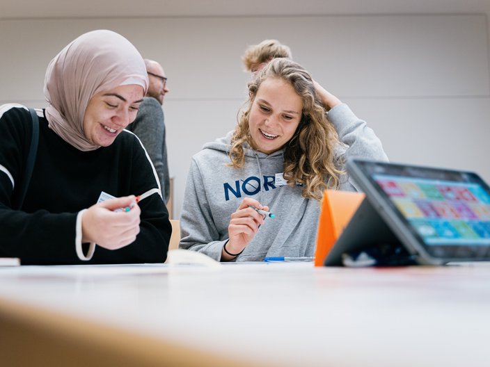 Zwei Schülerinnen sitzen an einem Tisch und erarbeiten etwas. Davor steht ein Ipad welches die SDGs anzeigt. Im Hintergrund unterhalten sich zwei Personen.