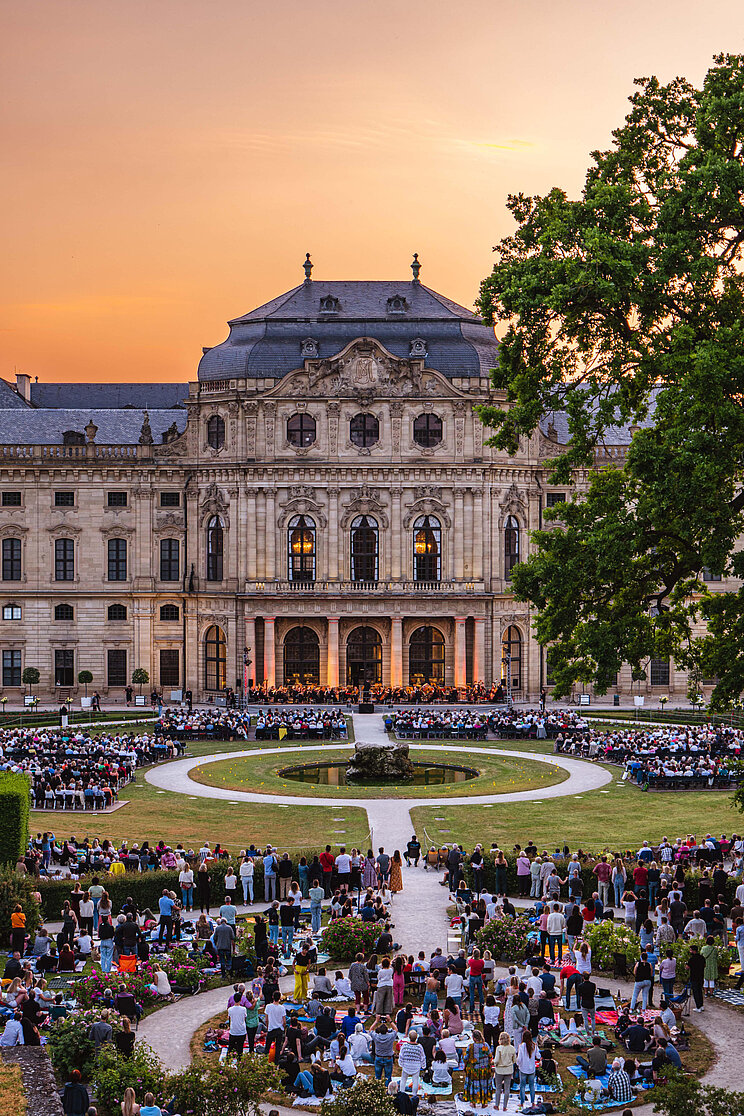 "Gemeinsamer Hörgenuss bei der Kleinen Nachtmusik". Das Foto der Residenz und des Hofgartens in Würzburg ist das Siegerbild 2024 des Fotowettbewerbs #WelterbeVerbindet Menschenmenge im Hofgarten an der Residenz in Würzburg
