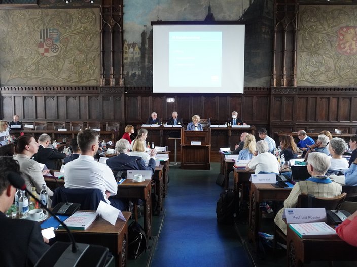 Blick in einen Plenarsaal. Im Hintergrund das Podium, an dem eine Frau zu den versammelten Menschen spricht.