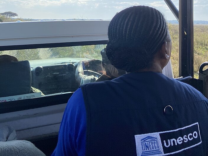 Eine Teilnehmerin in uNESCO-Weste ist von hinten abgebildet. Sie sitzt auf einem Auto, im Hintergrund erkennt man die langläufige Landschaft des Nationalparks. 
