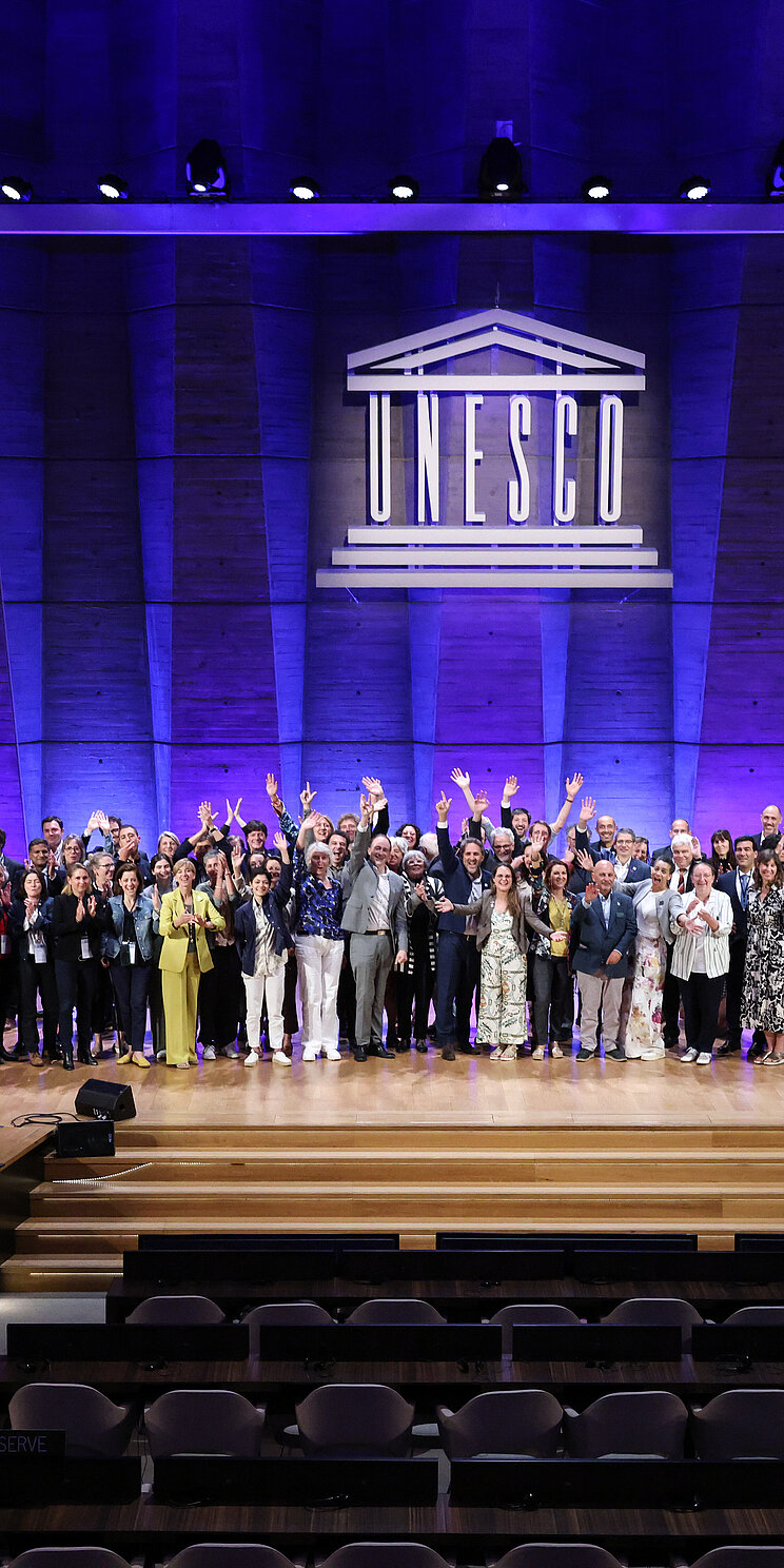Eine große Gruppe von Menschen posiert im Plenarsaal der UNESCO auf der Bühne. Im Hintergrund ist an der Wand das Logo der UNESCO angebracht.