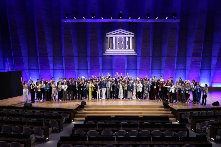 UNESCO HQ_Innenansicht Eine große Gruppe von Menschen posiert im Plenarsaal der UNESCO auf der Bühne. Im Hintergrund ist an der Wand das Logo der UNESCO angebracht.
