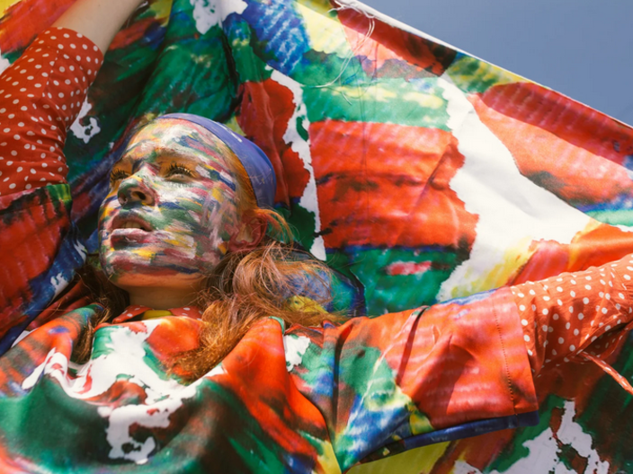 Das Bild zeigt eine junge Frau aus der unteren Ansicht, die eine bunt gemußterte Flagge hinter ihrem Rücken in die Höhe hält. Ihre Kleidung und ihr Gesicht sind mit den selben Farben bedeckt. In der oberen Rechten Ecke ist ein Stück blauer Himmel.