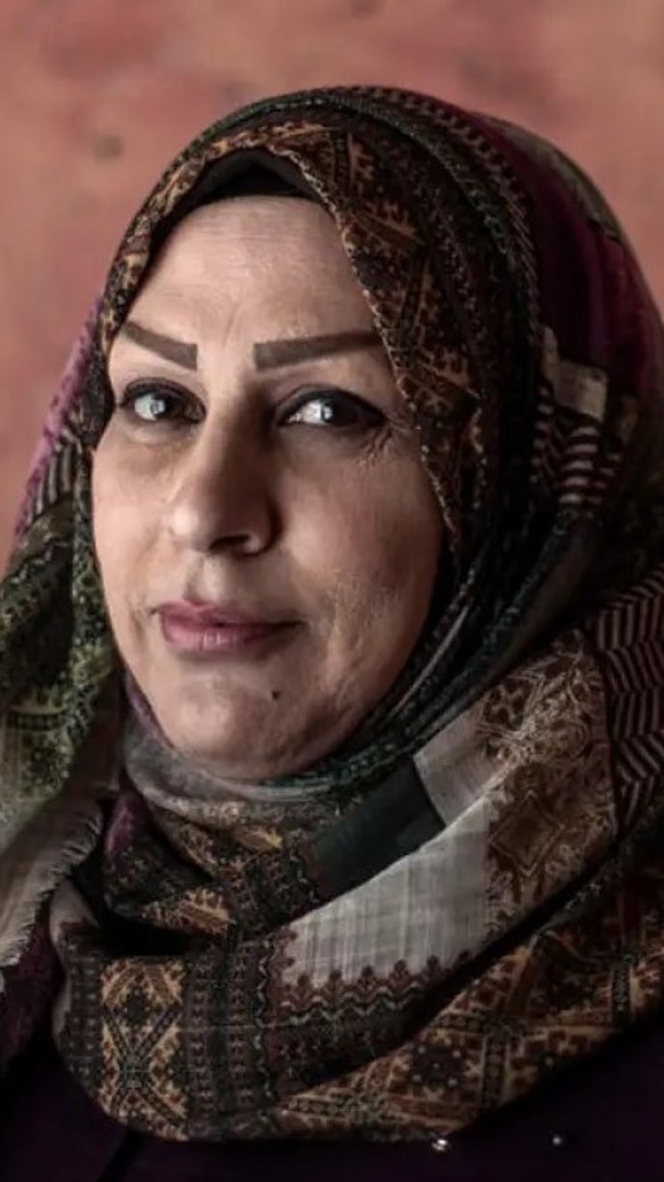 Eine Frau mit Kopftuch steht vor einer Mauer und schaut direkt in die Kamera