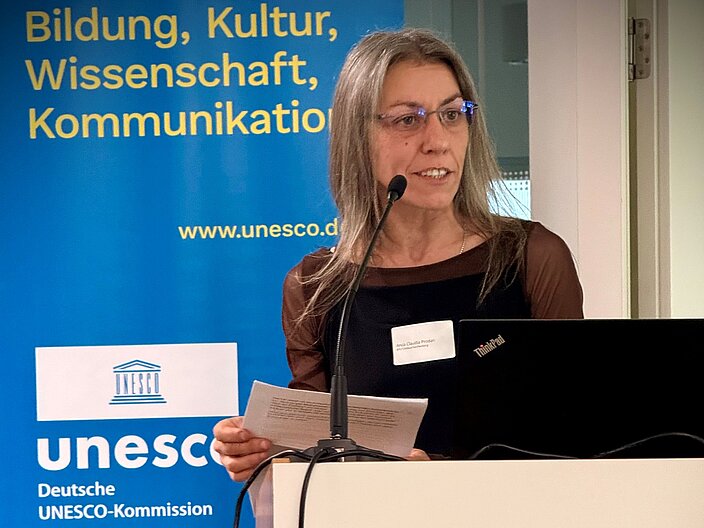 Anca Prodan steht an einem Redepult vor einem DUK-Banner und spricht in ein Mikrofon