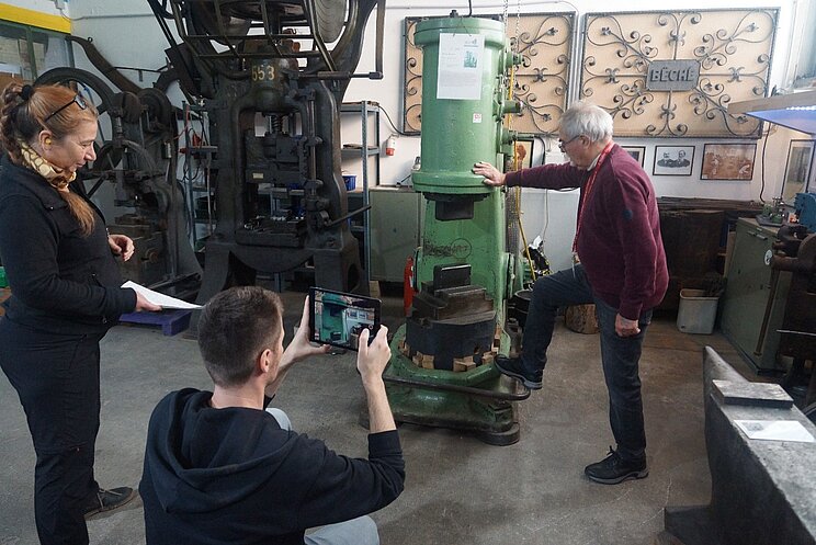 Drei Menschen stehen um eine gr&uuml;ne Fabrikmaschine herum. Ein Mann steht mit einem Fu&szlig; auf der Maschine, eine Frau mit einem Papier in der Hand schaut ihm dabei zu. Die dritte Person filmt Mann mit Maschine.