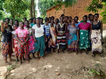 Die Gruppe der an dem Projekt teilnehmenden Frauen steht vor einem Haus im Dorf und schaut in die Kamera. 