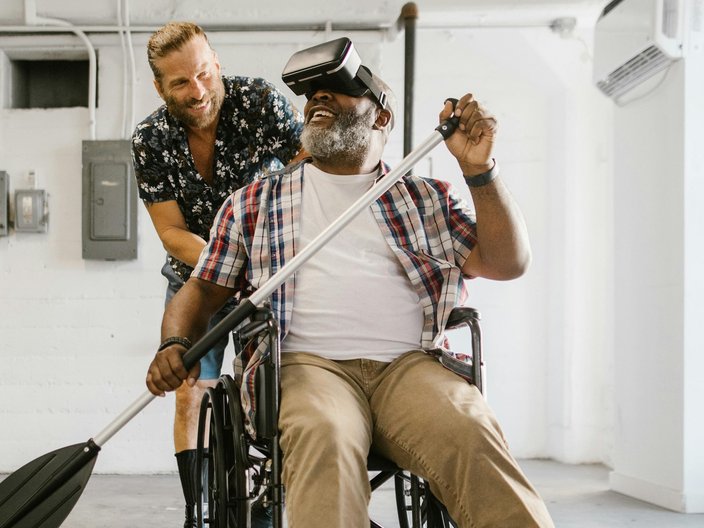 Ein älterer Mann mit dunkler Hautfarbe sitzt im Rollstuhl, trägt eine VR-Brille und hält ein Ruder in der Hand. Hinter ihm steht ein jüngerer Mann, der ihm vermutlich etwas erläutert. Beide Männer sehen fröhlich aus.