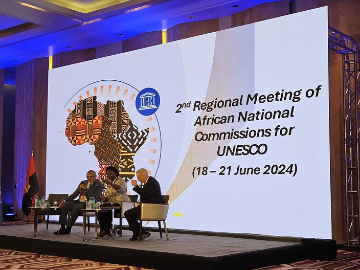 Three people sit on a stage and speak in a panel. Behind them is a large screen with the title ‘Second Regional Meeting of African National Commissions’