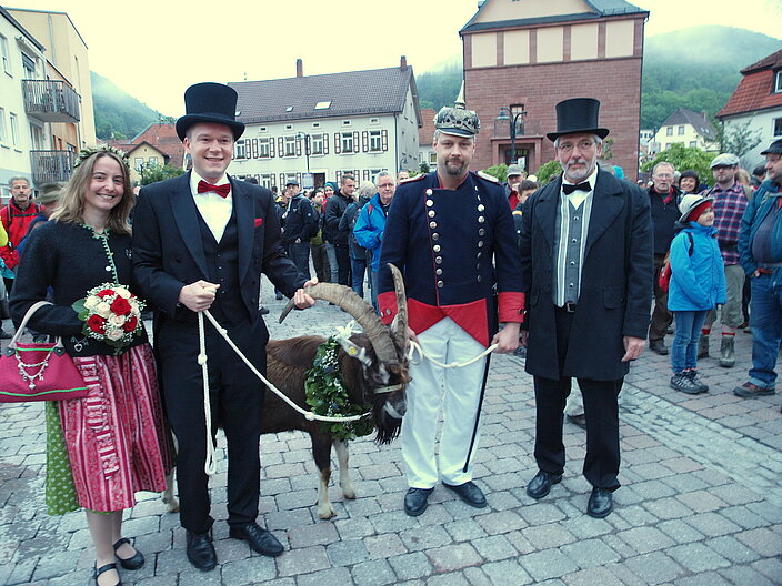 Zwei Männer in Sacko mit Zylinder und Militäroutfit führen einen Geißbock an zwei Seilen in der Mitte auf einem Dorfplatz in Lambrecht.