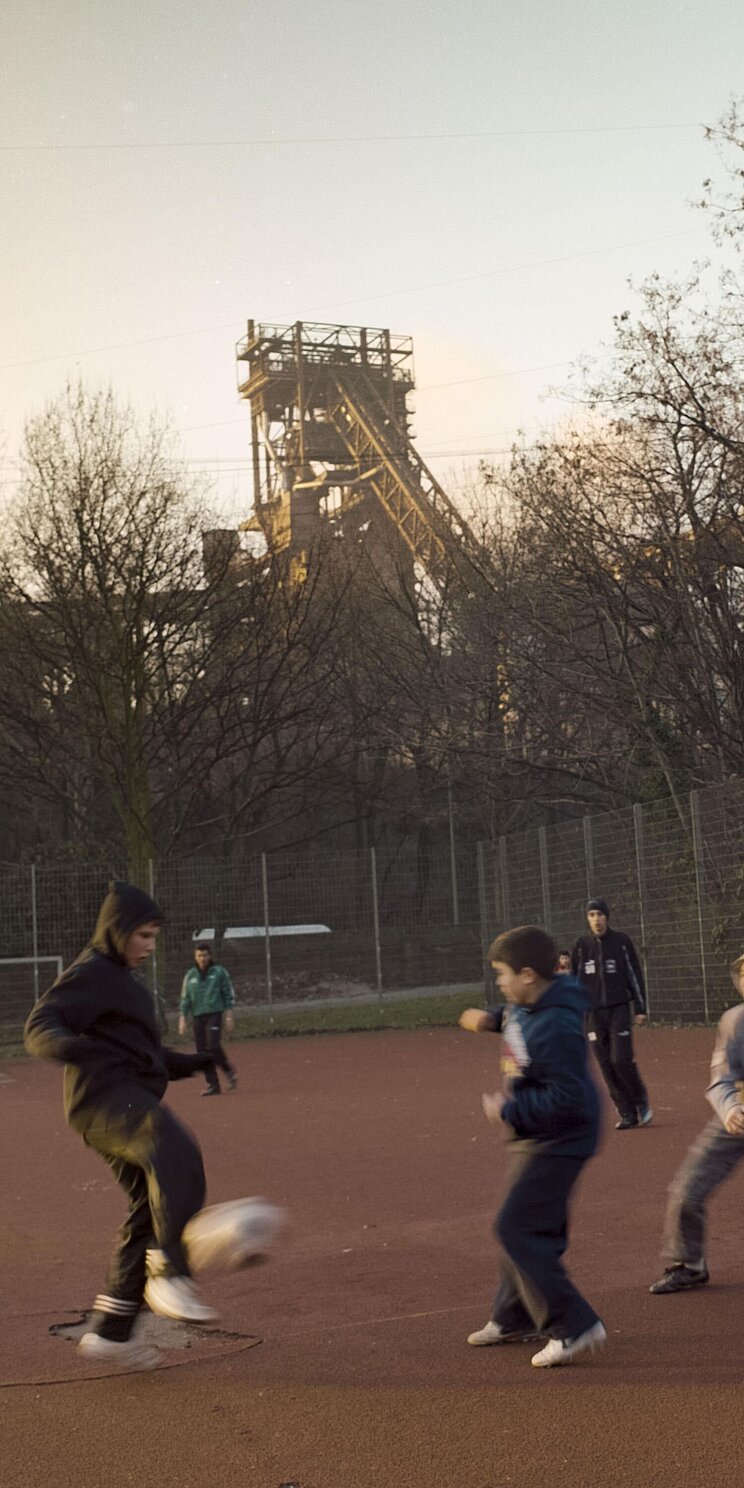 Sonneruntergang auf einem Bolzplatz mit Gummifl&auml;che. Kinder im Grundschulalter nutzen die Fl&auml;che zum Fu&szlig;ball spielen. Im Vordergrund versucht ein Kind den Ball zu verteidigen