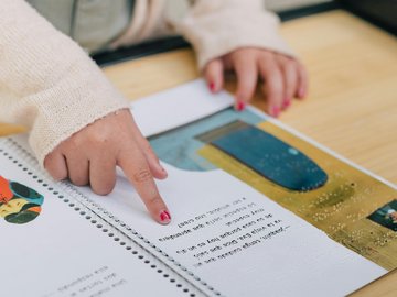 Mädchen liest Buch mit Brailleschrift Man sieht ein Kinderbuch mit Text, Brailleschrift und gezeichneten Bildern. Ein Finger fährt über die Buchseiten. An dem Finger sieht man abgeblätterten Nagellack. Die Hand sieht aus, als gehöre sie zu einem Mädchen im Grundschulalter.