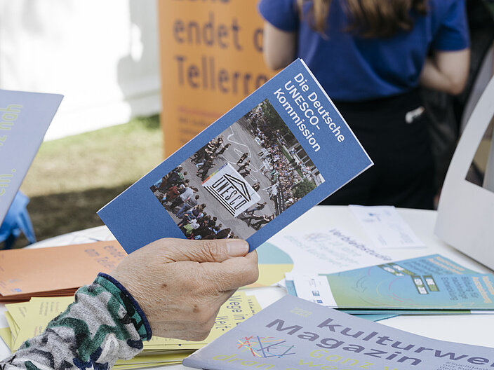 Eine Frauenhand hält einen länglichen blauen Flyer in der Hand. Im Hintergrund ist ein Tisch zu erkennen, auf dem weitere bunter Flyer liegen.
