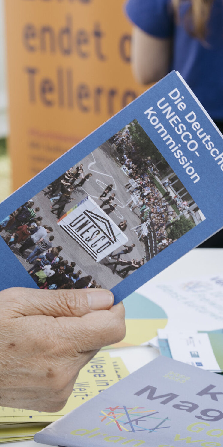 Eine Frauenhand hält einen länglichen blauen Flyer in der Hand. Im Hintergrund ist ein Tisch zu erkennen, auf dem weitere bunter Flyer liegen.