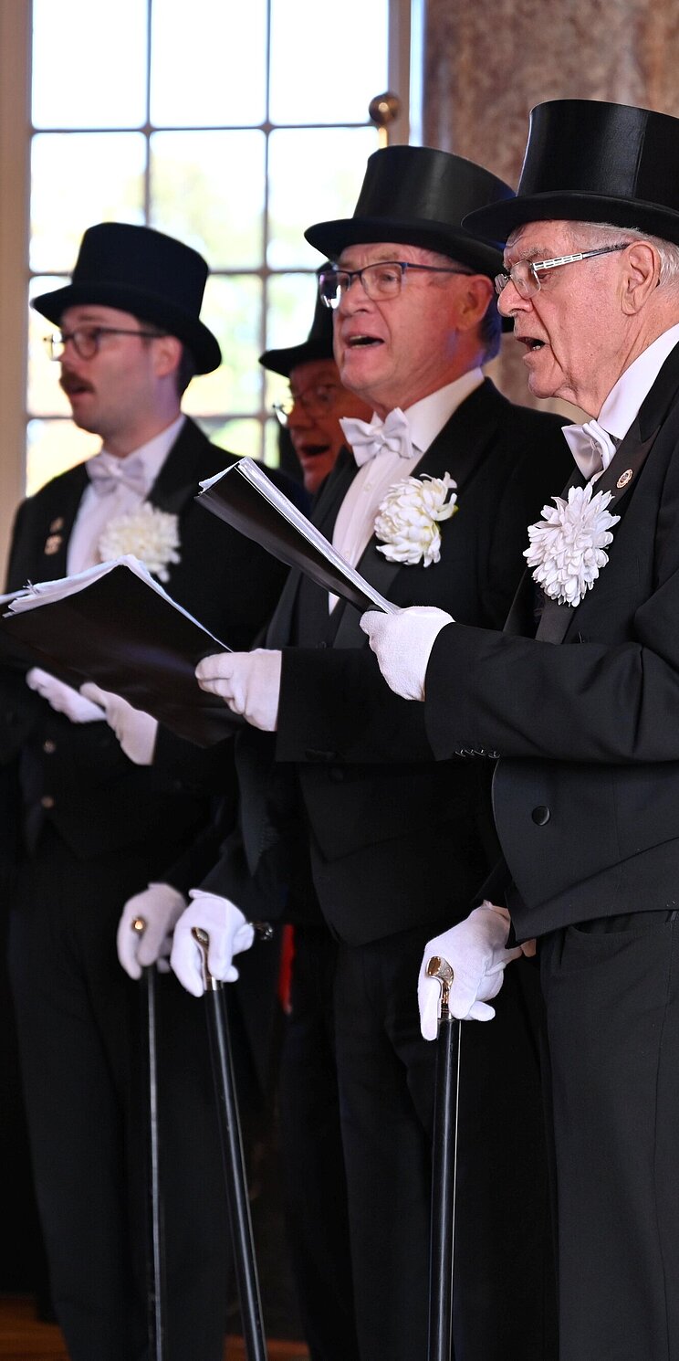 Finsterwalder_Sangestradition_Urkundenverleihing_2024 Vier Personen singen auf einer Bühne. Sie tragen schwarze Fräcke und Zylinder sowie weiße Hemden, Fliegen, Handschuhe und Blumen am Revers.