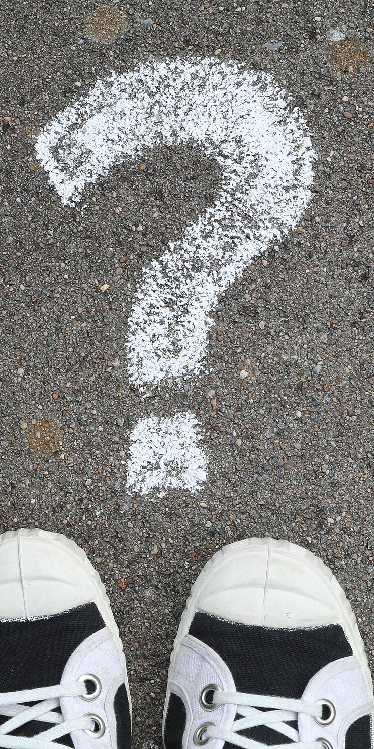 Fragezeichen Ein mit Kreide aufgemaltes Fragezeichen auf dem Boden. Unterhalb des Punktes sind zwei Schuhe zu sehen.