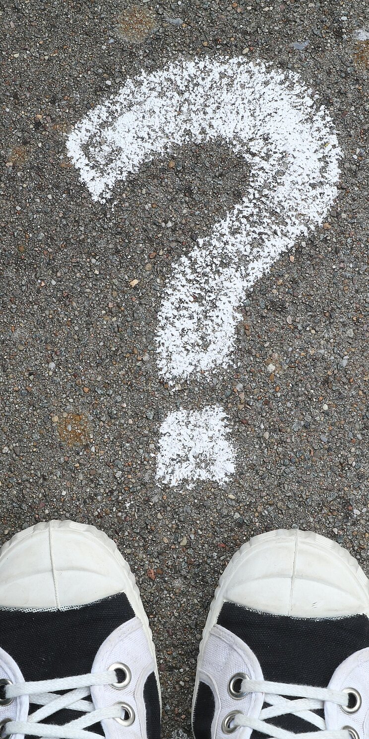 Ein mit Kreide aufgemaltes Fragezeichen auf dem Boden. Unterhalb des Punktes sind zwei Schuhe zu sehen.