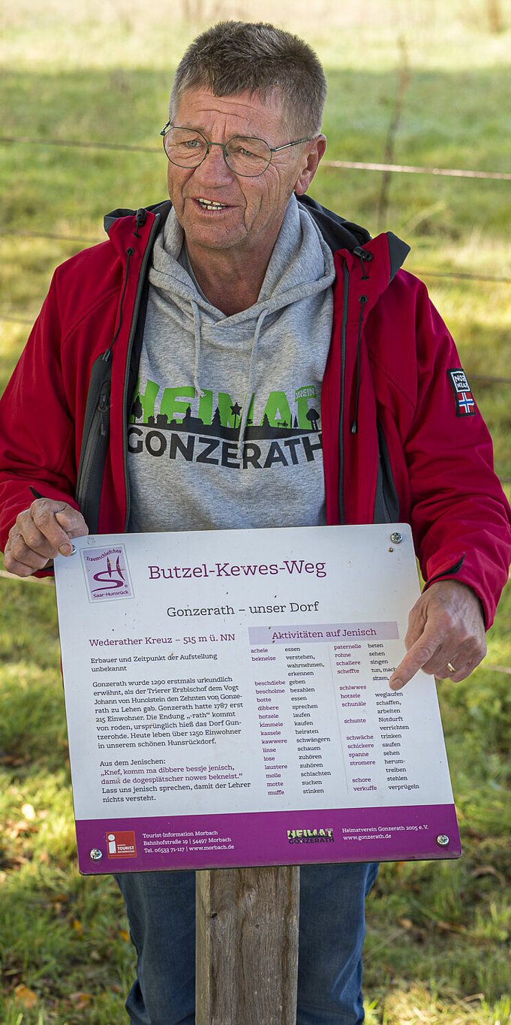 07_Rotwelsch-Dialekte_Butzel-Kewes-Weg__Sprechtafel_c_IGS Eine Person hält eine Sprechtafel auf dem Butzel Kewes Weg in die Höhe und erklärt die Begriffe einer zweiten Person.