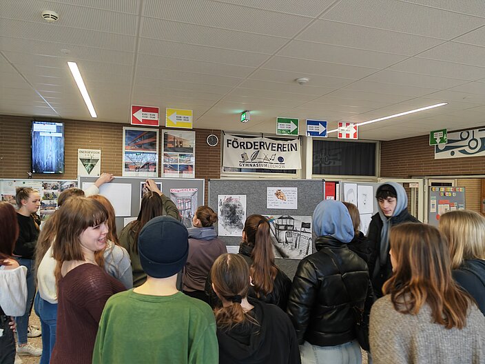 Schüler*innen stehen vor Pinnwänden in der Schule auf denen die Eindrücke in Schrift und Bild von dem KZ Besuch ausgestellt sind.