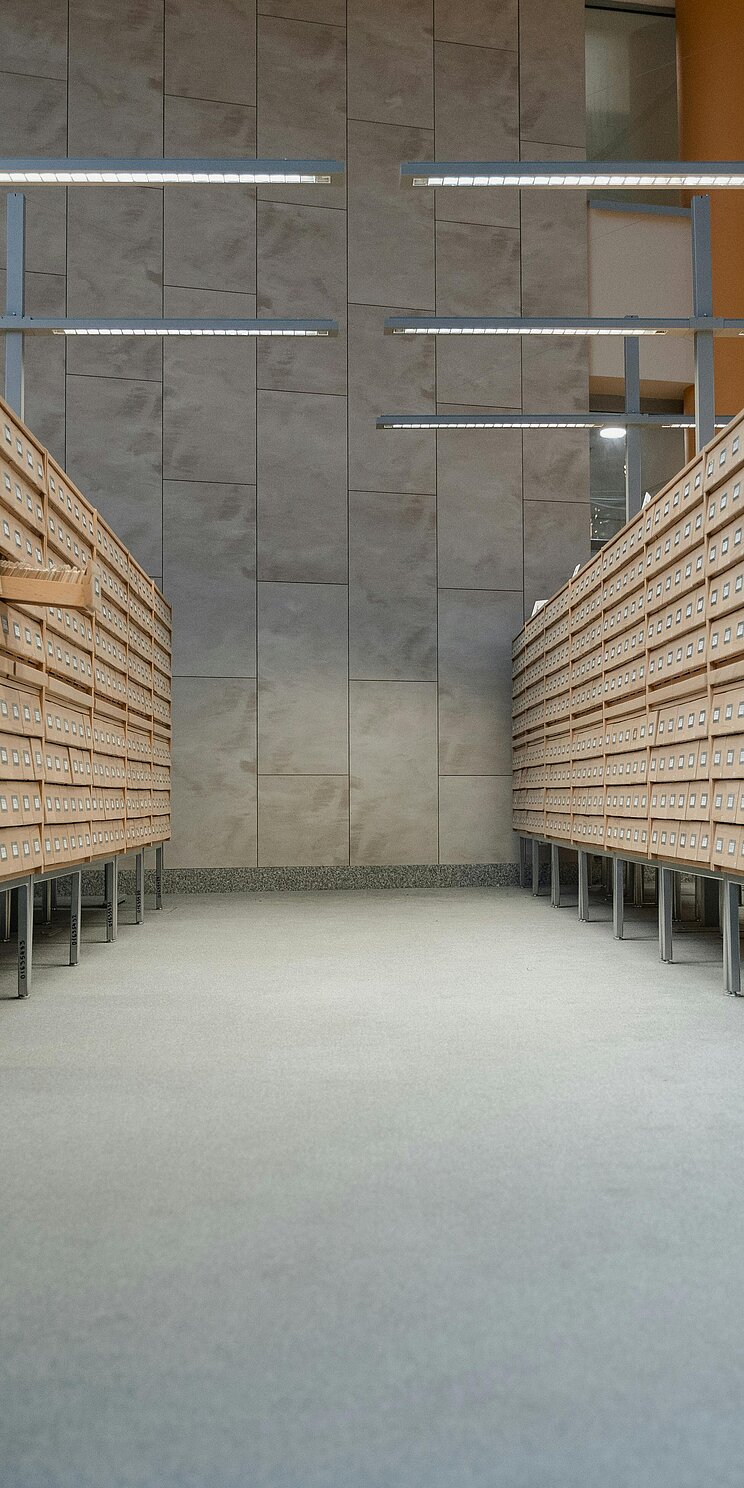 Zu beiden Seiten sind Archivregale mit Schubladen zusehen. In der Mitte ein grauer Gang.