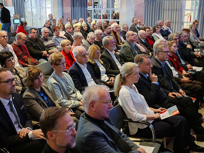 Urkundenverleihung_25_Publikum Perspektive auf das Publikum. Mehrere Stuhlreihen hintereinander voll besetzt.