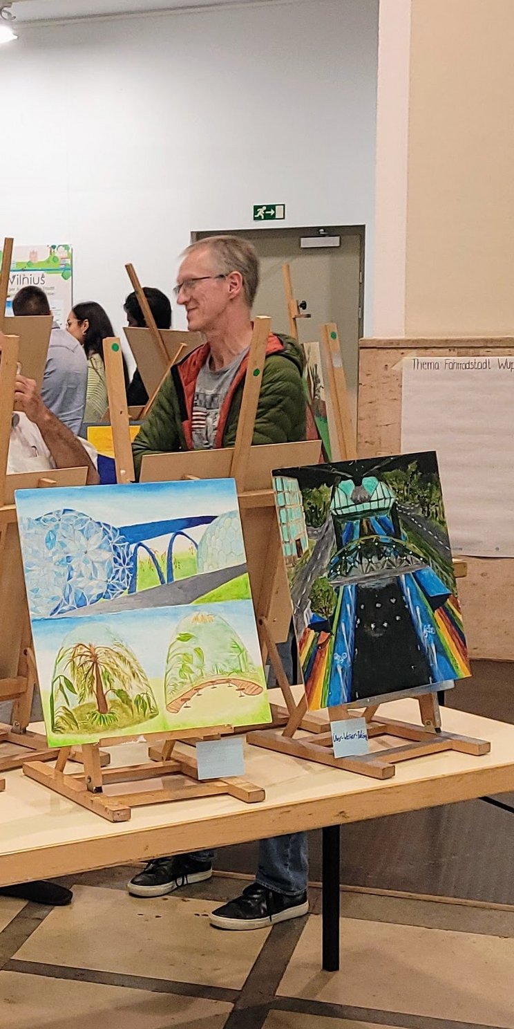 Ein Gallerywalk mit Kunstbildern von Schülern und Schülerinnen des Ganztagsgymnasium Johannes Rau. Die Bilder sind auf Leinwände gemalt, stehen auf Staffeln auf Tischen und mehrere Menschen schauen sich die Ausstellung an. Eine Leinwand zeigt eine Weltkugel und ein andere ein Stadt und ein Naturmotiv.