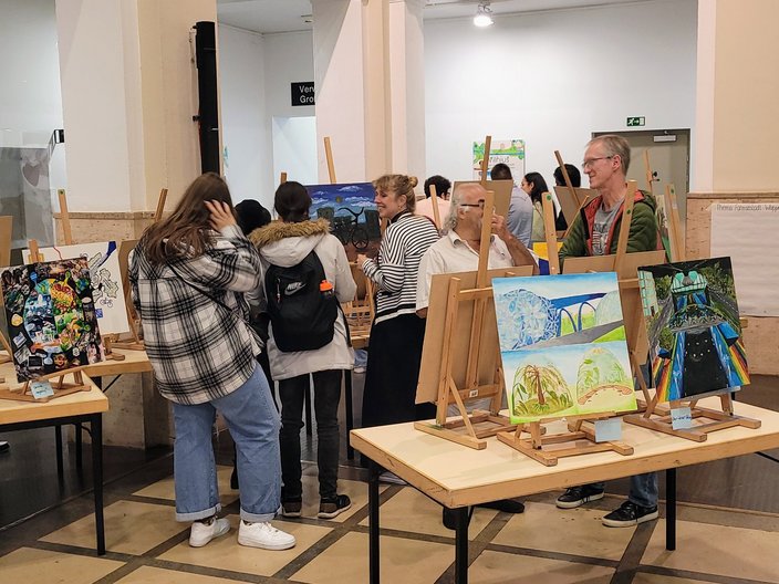 Ein Gallerywalk mit Kunstbildern von Schülern und Schülerinnen des Ganztagsgymnasium Johannes Rau. Die Bilder sind auf Leinwände gemalt, stehen auf Staffeln auf Tischen und mehrere Menschen schauen sich die Ausstellung an. Eine Leinwand zeigt eine Weltkugel und ein andere ein Stadt und ein Naturmotiv.
