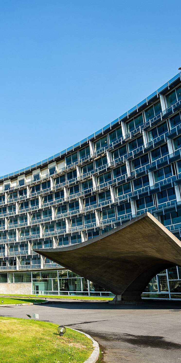 UNESCO Headquarter Blick auf ein konkave Gebäudefront mit expressiven Eingang aus Beton- Auf der linke Bildhälfte eine abstrakte Skulptur aus Stahl.
