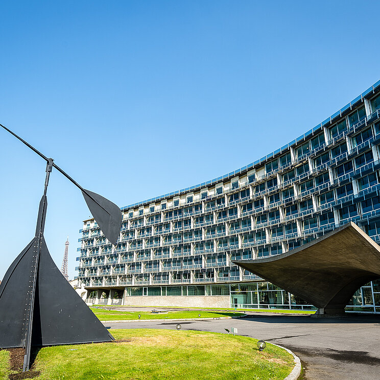 UNESCO Headquarter Blick auf ein konkave Gebäudefront mit expressiven Eingang aus Beton- Auf der linke Bildhälfte eine abstrakte Skulptur aus Stahl.