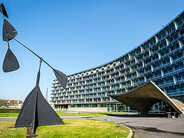 Blick auf ein konkave Geb&auml;udefront mit expressiven Eingang aus Beton- Auf der linke Bildh&auml;lfte eine abstrakte Skulptur aus Stahl.