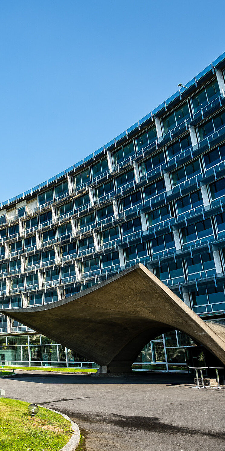 Blick auf ein konkave Gebäudefront mit expressiven Eingang aus Beton- Auf der linke Bildhälfte eine abstrakte Skulptur aus Stahl.