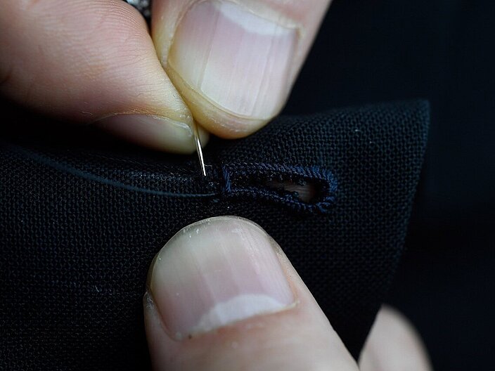 Nahaufnahme, auf der zwei Daumen zu sehen sind und ein Zeigefinger. Mit einer kleinen Nadel wird ein Knopfloch in den Stoff gestochen