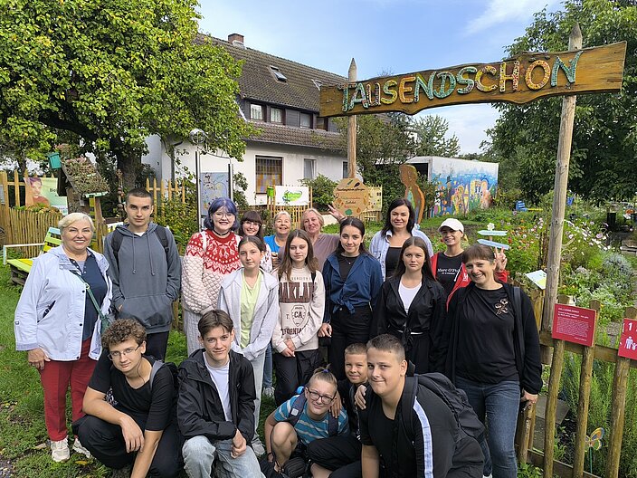 Die Gruppe von ukrainischen Teilnehmenden zusammen mit den Lehrkräften steht vor dem Eingang des Gartens Tausendschön. Über dem Eingang hängt ein Holzschild mit dem Namen des Gartens.