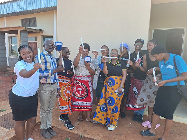 Eswatini_2024_Kerzenproduktion 10 Jugendliche stehen gesammelt als Gruppe vor einer beigen Wand und halten ihre selbst hergestellten Kerzen lächelnd in die Kamera.