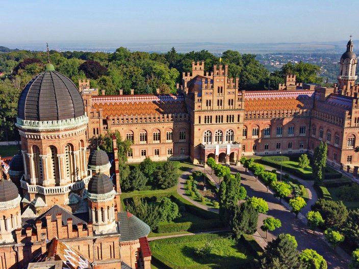 Blick von oben auf ein Residenzensemble mit vielen Türmen, Verzierungen und dunklen Kuppeldächern. Der Innenhof ist bepflanzt und gut gepflegt. Im Hintergrund sieht man einen grünen Hügel und eine Stadt von oben.