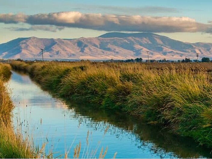 Man sieht mittig-links einen Fluss der in die Ferne geht und daneben eine Wiese in gelb-braunen Farbtönen. Im Hintergrund schaut man auf die Berge. Die Farben am Himmel lassen einen Sonnenuntergang vermuten.
