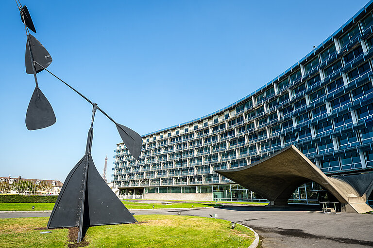 Blick auf das Geb&auml;ude der UNESCO in Paris. Ein mehrst&ouml;ckiges Geb&auml;ude mit vielen Fenstern, davor ein Garten mit Skulpturen, im Hintergrund der Eifelturm.