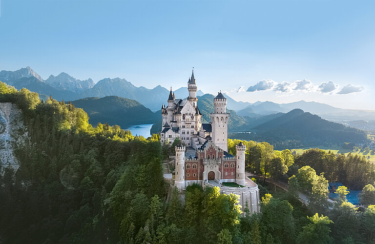 Blick auf das Schloss Neuschwanstein vom Osten aus - auf einem Berg zwischen Bäumen, im Hintergeund ein See und das Alpenpanorama