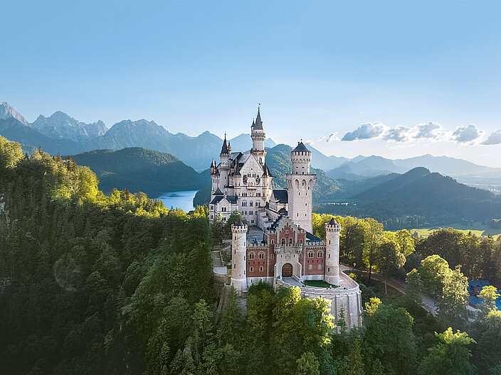 Blick auf das Schloss Neuschwanstein vom Osten aus - auf einem Berg zwischen Bäumen, im Hintergeund ein See und das Alpenpanorama
