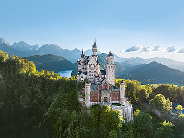 Blick auf das Schloss Neuschwanstein vom Osten aus - auf einem Berg zwischen Bäumen, im Hintergeund ein See und das Alpenpanorama