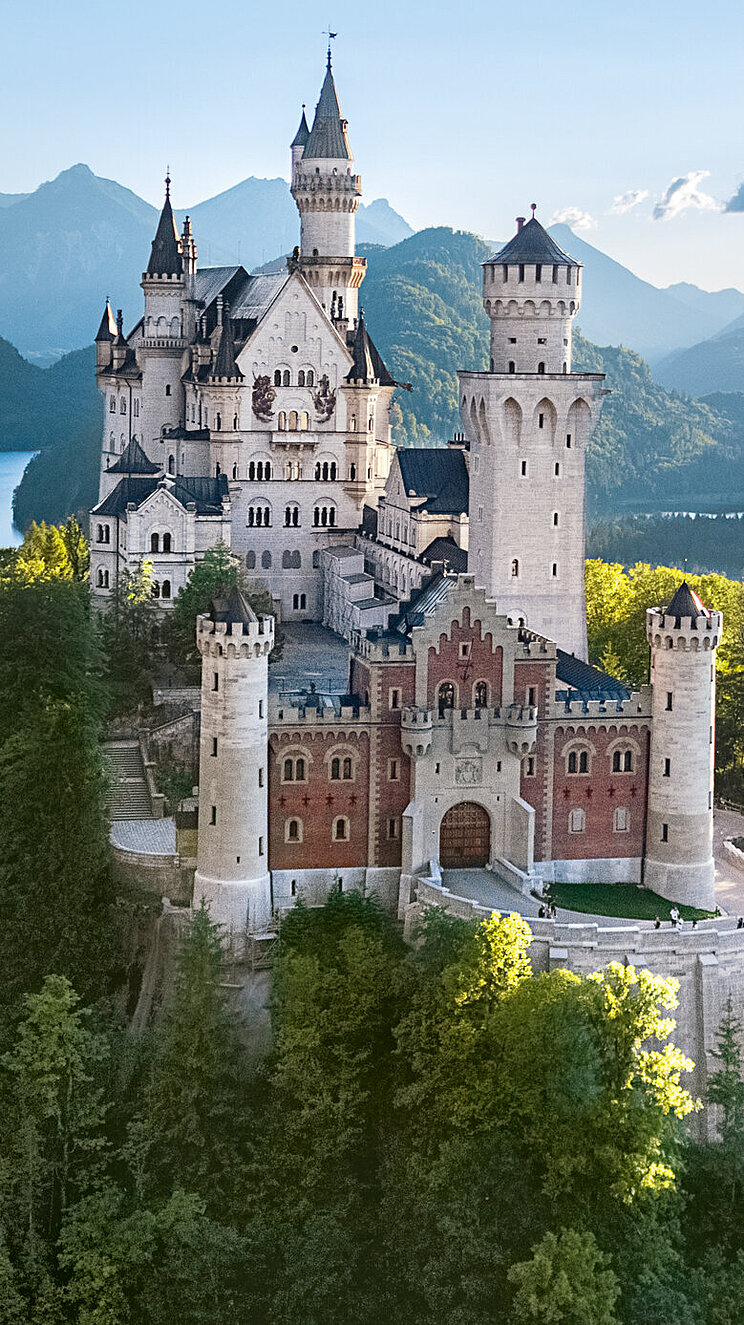 Blick auf das Schloss Neuschwanstein vom Osten aus - auf einem Berg zwischen Bäumen, im Hintergeund ein See und das Alpenpanorama
