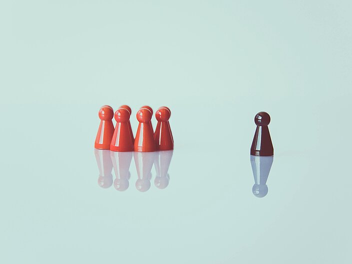 Vor hellem Hintergrund stehen vier rote Spielfiguren auf der linken Seite. Rechts mit etwas Abstand steht eine schwarze Spielfigur.